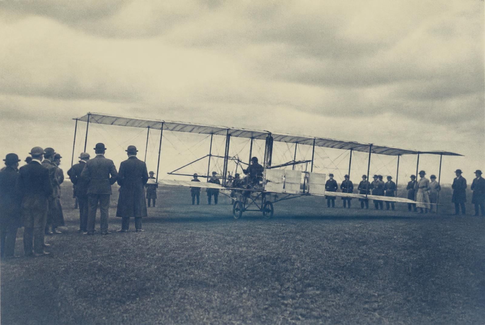 An early biplane on a grass field with period spectators